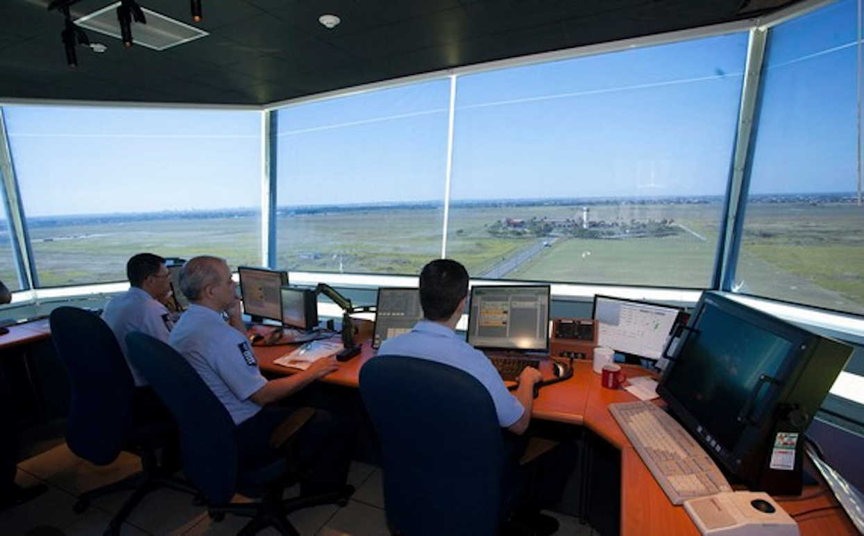 Una vista panorámica de una torre de control blanca y alta rodeada de aviones y pistas de aterrizaje